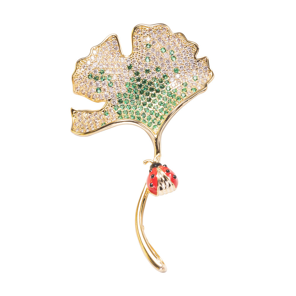 A brooch pin featuring a gold-colored ginkgo leaf design with green and red accents and a yellow gold over brass base.