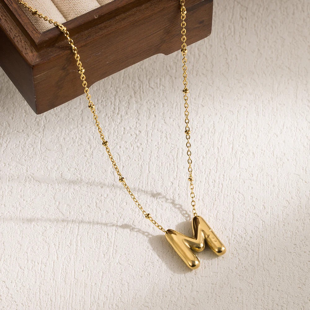 A gold-colored stainless steel pendant necklace with an 'M' initial design, displayed against a wooden backdrop.