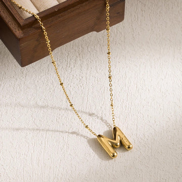 A gold-colored stainless steel pendant necklace with an 'M' initial design, displayed against a wooden backdrop.