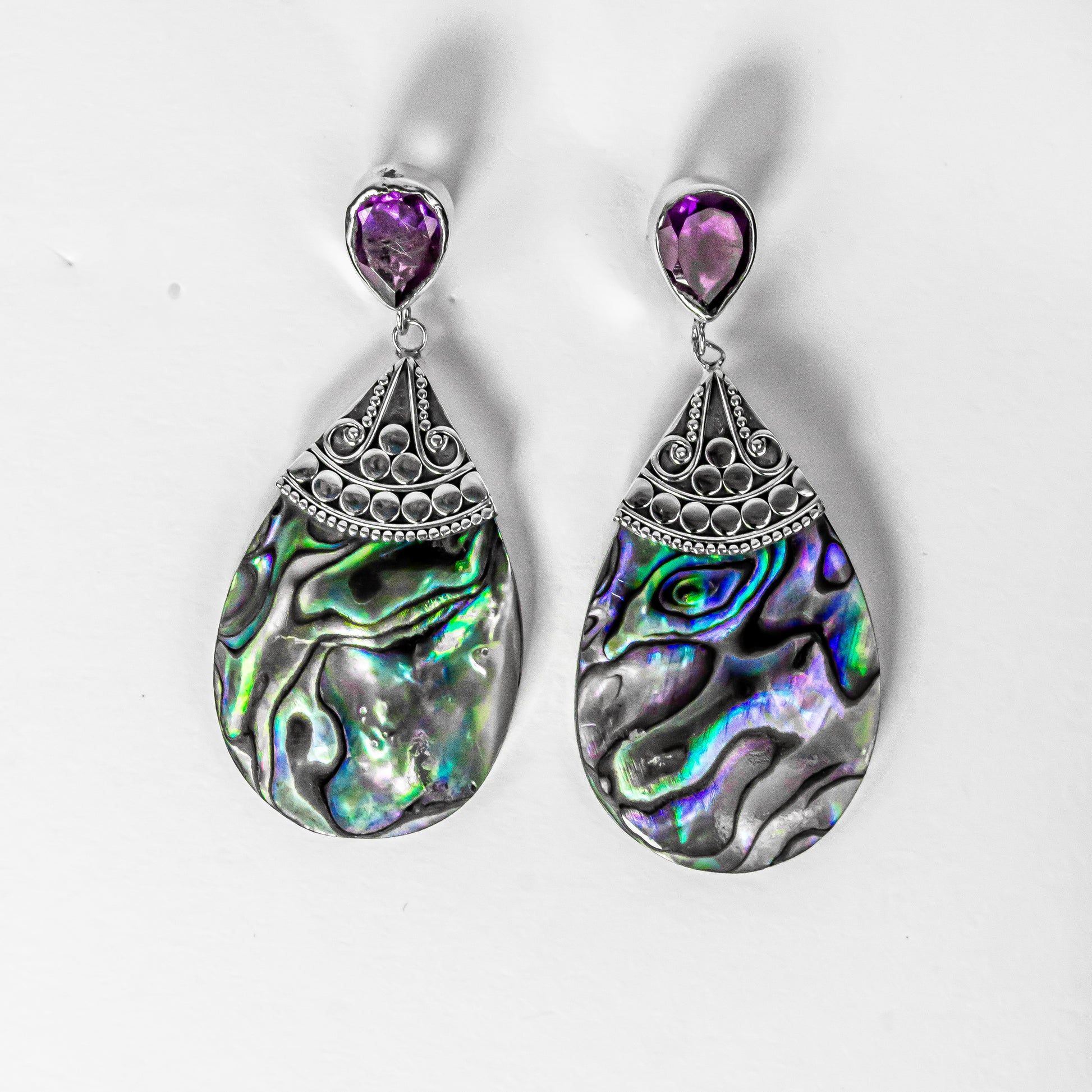 A pair of sterling silver earrings with abalone shell and amethyst gemstone accents, featuring intricate traditional Balinese motifs.