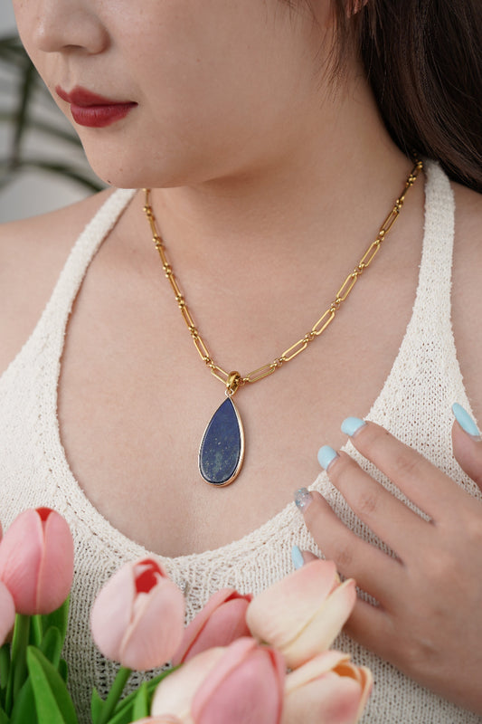 Woman wearing a gold necklace with a blue lapis pendant, surrounded by pink tulips.