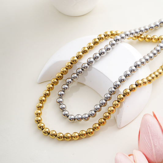 Two beaded necklaces, one gold and one silver, on a white surface with a pink flower.