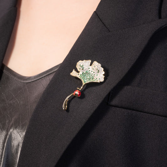 A brooch pin featuring a gold-colored ginkgo leaf design with green and red accents and a yellow gold over brass base.